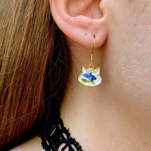 Puede incluir: Pendientes dorados con forma de gato y acabado esmaltado blanco. Cada pendiente presenta un dise&ntilde;o de flor azul. Los pendientes est&aacute;n suspendidos de ganchos dorados. Un accesorio &uacute;nico.