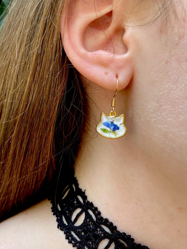 Puede incluir: Pendientes dorados con forma de gato y acabado esmaltado blanco. Cada pendiente presenta un dise&ntilde;o de flor azul. Los pendientes est&aacute;n suspendidos de ganchos dorados. Un accesorio &uacute;nico.