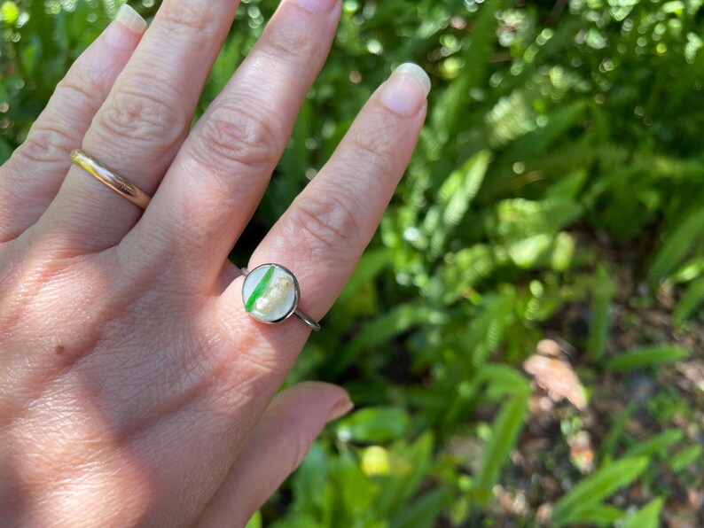 May include: A silver ring with a round, clear resin setting featuring a green leaf and white flowers.