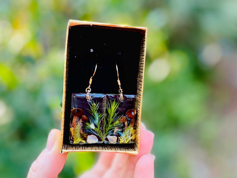 May include: A pair of square resin earrings with gold hooks. The earrings feature a pressed botanical design with small mushrooms, green moss, and a sprig of pine needles.