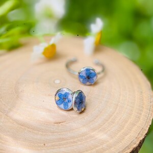 K&ouml;nnte beinhalten: Zwei silberne Ringe mit blauen Blumenakzenten. Die Ringe liegen auf einer Holzoberfl&auml;che.