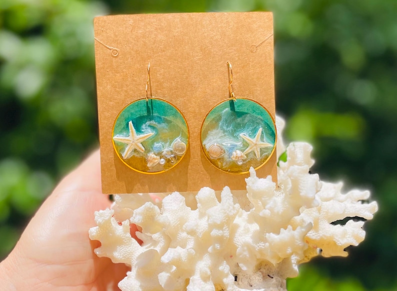 Op de afbeelding: Goudkleurige, ronde oorbellen met een strandthema. Elke oorbel heeft een witte zeester, kleine schelpen en een turquoise en witte harsachtergrond. De oorbellen worden gepresenteerd op een bruine kaart.