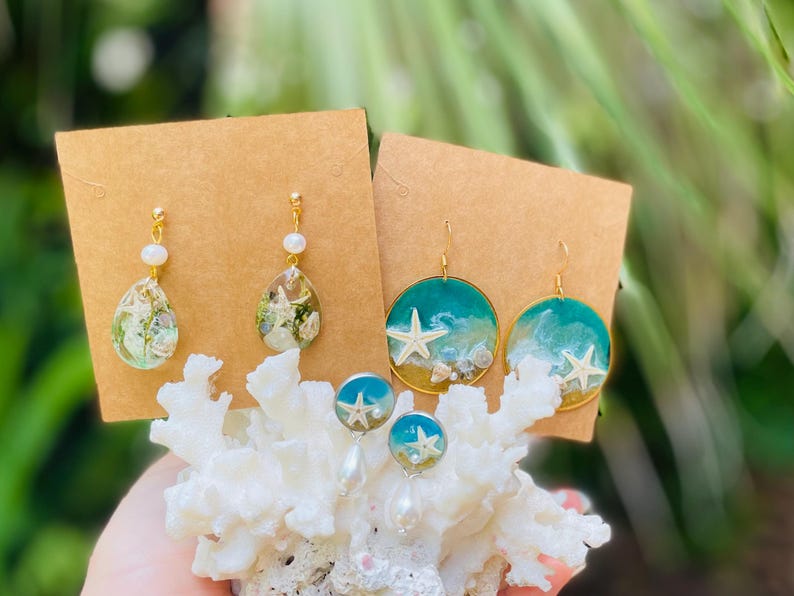 Op de afbeelding: Een collectie handgemaakte oorbellen met oceaan-thema ontwerpen. De oorbellen bevatten druppelvormige harsstukken met ingebedde schelpen en parels, en ronde oorbellen met zeesterren en oceaangolfontwerpen. De oorbellen worden gepresenteerd op bruine kaarten.