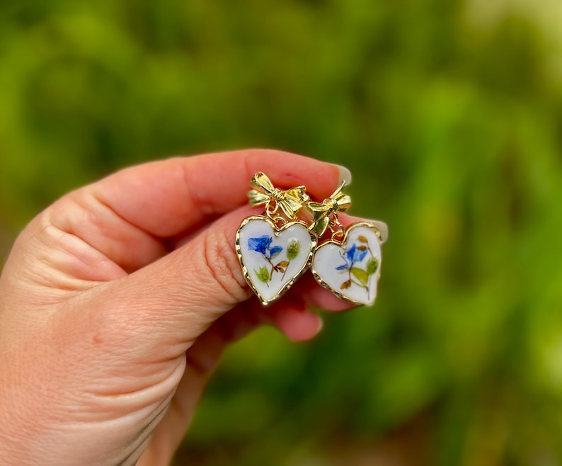 May include: Two gold heart-shaped earrings with a white background and blue and green pressed flowers inside. The earrings have a gold bow at the top.