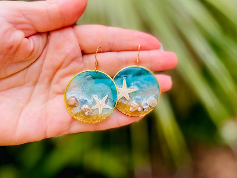 Op de afbeelding: Een paar ronde, goudkleurige oorbellen. Elke oorbel heeft een miniatuur strandsc&egrave;ne met een witte zeester, schelpen en een turquoise en wit oceaanontwerp. De oorbellen worden in een hand gehouden tegen een vage groene achtergrond.