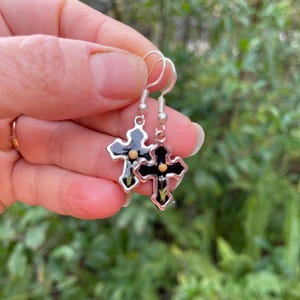 May include: A pair of silver dangle earrings with black cross charms. Each cross has a small gold dot in the center.