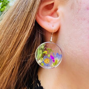 Puede incluir: Pendientes circulares transparentes con flores secas encapsuladas en resina. Los pendientes presentan una variedad de flores coloridas, incluyendo azules, amarillas y moradas, sobre un fondo transparente. Cuelgan de ganchos plateados.