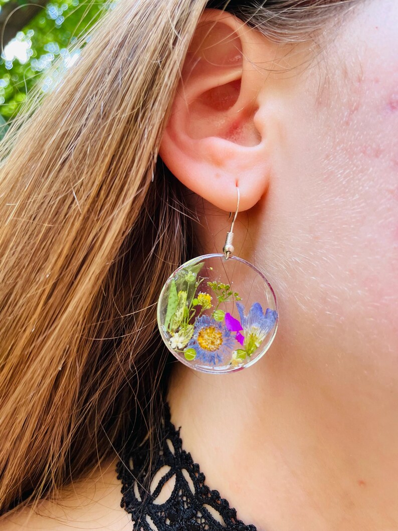 Puede incluir: Pendientes circulares transparentes con flores secas encapsuladas en resina. Los pendientes presentan una variedad de flores coloridas, incluyendo azules, amarillas y moradas, sobre un fondo transparente. Cuelgan de ganchos plateados.