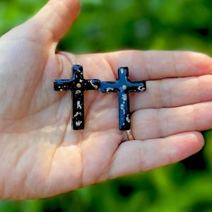 May include: A pair of black cross earrings with gold accents. The crosses are made of resin and have a speckled finish.