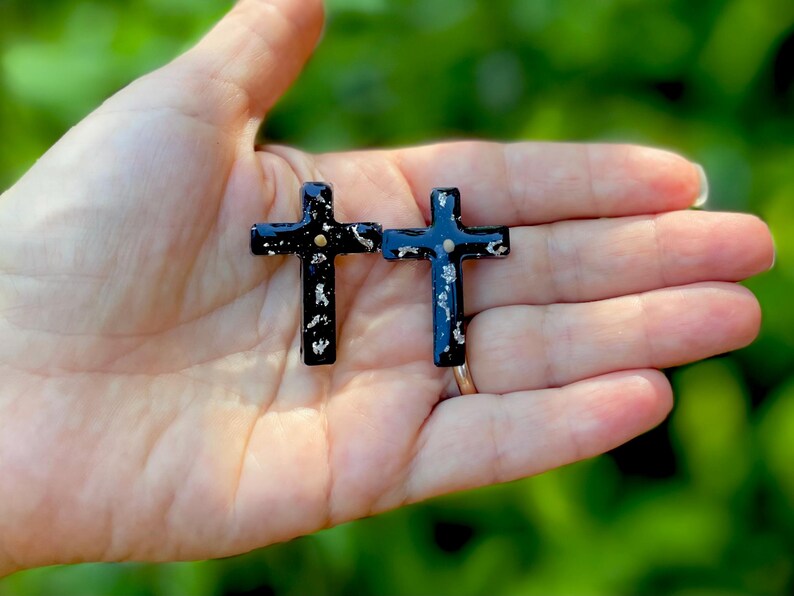 May include: A pair of black cross earrings with gold accents. The crosses are made of resin and have a speckled finish.
