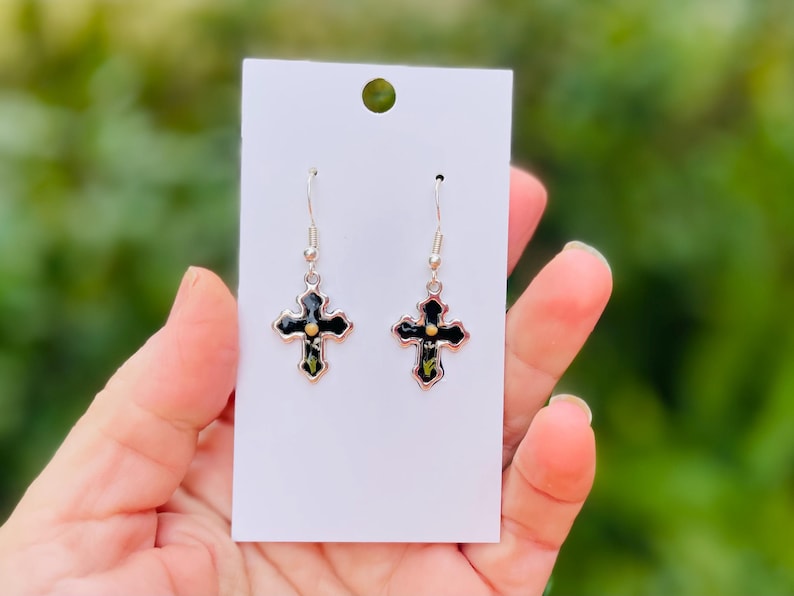 May include: A pair of silver dangle earrings with black cross charms. Each cross has a small gold dot in the center.