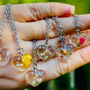 Puede incluir: Cinco collares de plata con colgantes en forma de coraz&oacute;n transparentes llenos de flores y vegetaci&oacute;n prensadas. Cada colgante tiene una disposici&oacute;n diferente de flores y vegetaci&oacute;n.