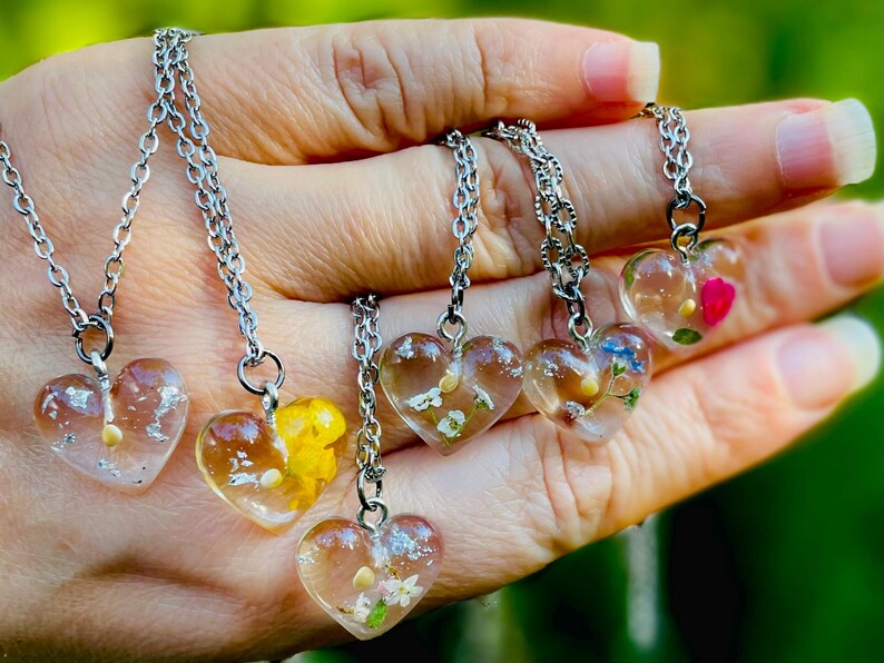 Puede incluir: Cinco collares de plata con colgantes en forma de coraz&oacute;n transparentes llenos de flores y vegetaci&oacute;n prensadas. Cada colgante tiene una disposici&oacute;n diferente de flores y vegetaci&oacute;n.