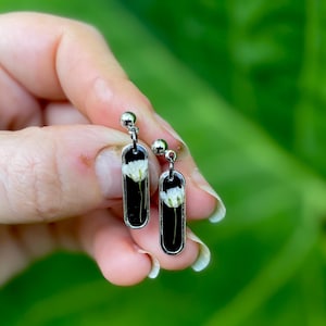 Puede incluir: Pendientes colgantes plateados con fondo negro y una flor blanca encapsulada en resina. Los pendientes tienen un pequeño perno redondo en la parte superior. El fondo es verde borroso.