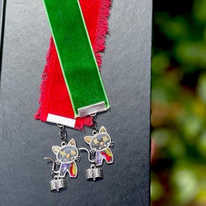 May include: Two green and red velvet ribbon bookmarks with silver cat charms. Each charm features a black cat with a book in its paws and a glittery design.