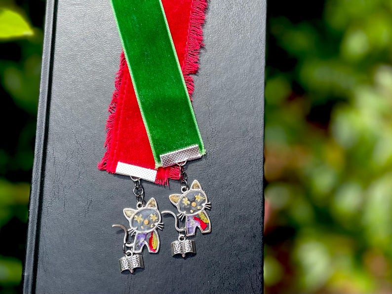 May include: Two green and red velvet ribbon bookmarks with silver cat charms. Each charm features a black cat with a book in its paws and a glittery design.