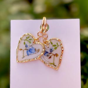 May include: A pair of gold heart-shaped earrings with a white background and blue and green pressed flowers inside.