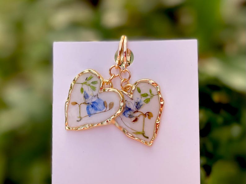 May include: A pair of gold heart-shaped earrings with a white background and blue and green pressed flowers inside.