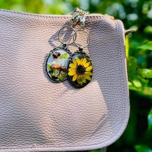 May include: Two oval keyrings with botanical designs hang from a silver chain. One features mushrooms, the other a yellow sunflower. The keyrings are attached to a light pink leather purse, with a blurred green background.