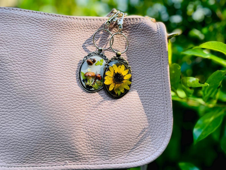 May include: Two oval keyrings with botanical designs hang from a silver chain. One features mushrooms, the other a yellow sunflower. The keyrings are attached to a light pink leather purse, with a blurred green background.
