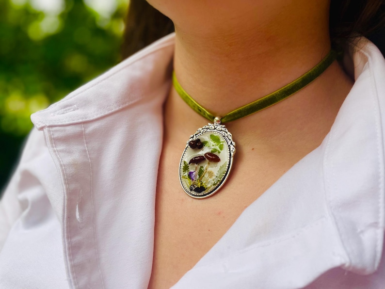 K&ouml;nnte beinhalten: Eine gr&uuml;ne Samt-Chokerkette mit einem silbernen ovalen Anh&auml;nger mit gepressten Blumen und Pilzen.