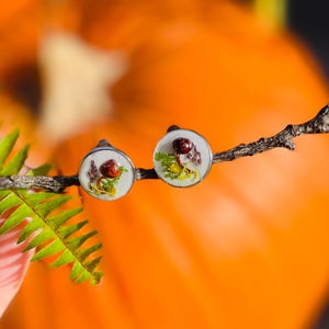 Puede incluir: Un par de pendientes de plata con fondo blanco y un dise&ntilde;o de flores prensadas. Los pendientes est&aacute;n en una rama con hojas verdes y una calabaza naranja borrosa en el fondo.