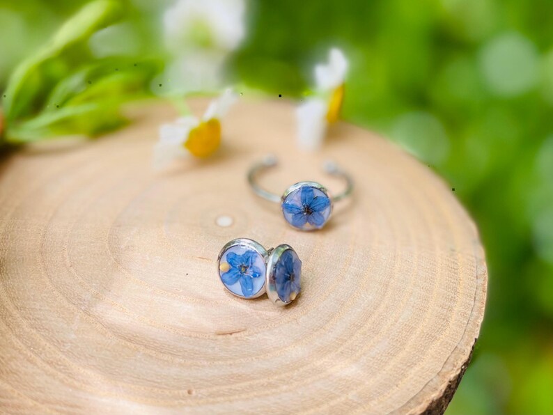 K&ouml;nnte beinhalten: Zwei silberne Ringe mit blauen Blumenakzenten. Die Ringe liegen auf einer Holzoberfl&auml;che.