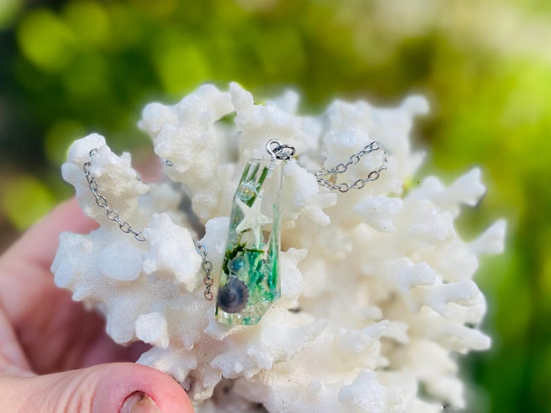 K&ouml;nnte beinhalten: Halskette mit Anh&auml;nger aus klarem Harz und einer silbernen Kette. Der Anh&auml;nger enth&auml;lt einen kleinen Seestern, gr&uuml;ne Algen und eine Muschel. Die Halskette wird auf einer wei&szlig;en, korallenartigen Struktur vor unscharfem Hintergrund pr&auml;sentiert.