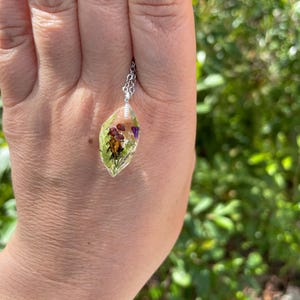 May include: A silver chain necklace with a clear resin pendant containing pressed flowers and leaves. The pendant is shaped like a teardrop and has a light green hue.