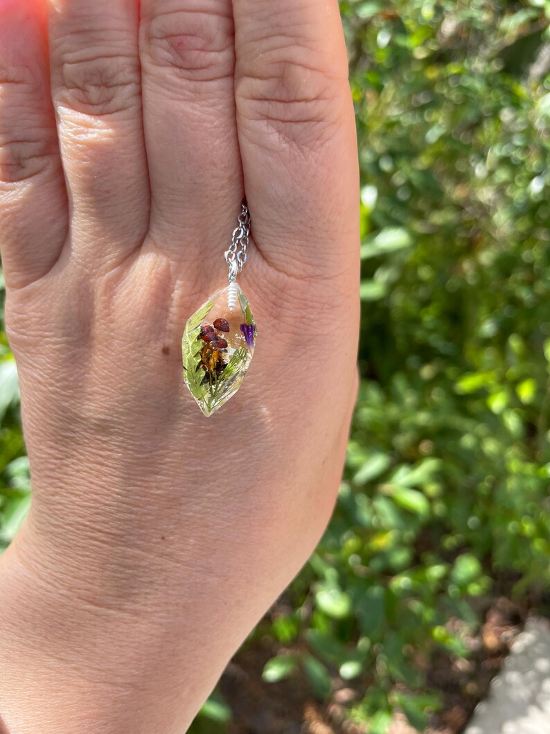 May include: A silver chain necklace with a clear resin pendant containing pressed flowers and leaves. The pendant is shaped like a teardrop and has a light green hue.