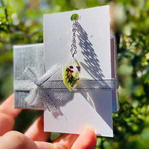 May include: A silver chain necklace with a clear teardrop pendant containing pressed flowers and greenery. The pendant is on a white card with a silver ribbon.