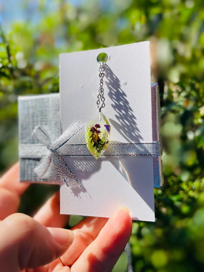 May include: A silver chain necklace with a clear teardrop pendant containing pressed flowers and greenery. The pendant is on a white card with a silver ribbon.