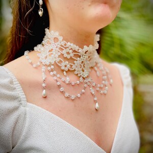 May include: A white beaded choker necklace with a delicate lace design and pearl accents. The necklace features multiple strands of beads and a teardrop-shaped pendant.