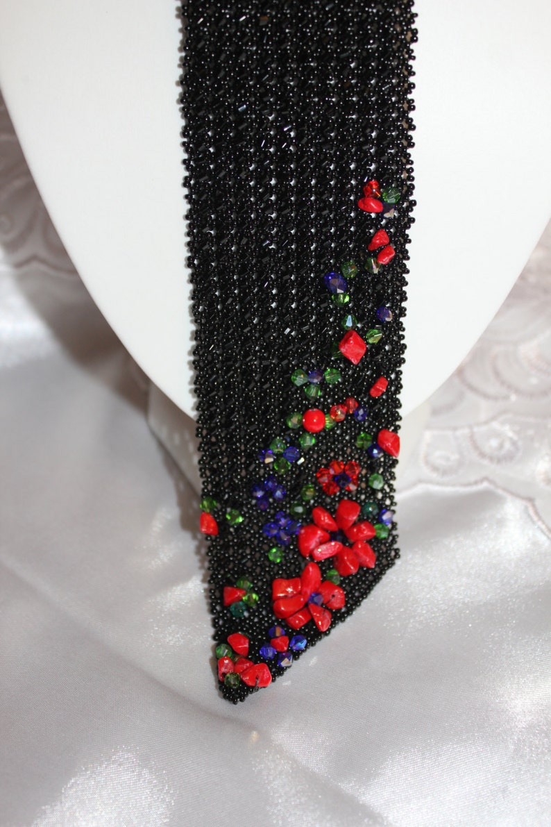 May include: A black beaded necklace with a floral design. The necklace features red, green, and blue gemstone accents. The necklace is displayed on a white mannequin head.