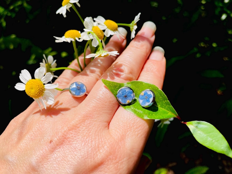 K&ouml;nnte beinhalten: Ein Set aus silbernen Ohrringen und einem Ring mit blauen Vergissmeinnicht-Bl&uuml;ten, die in klarem Harz gepresst sind. Der Schmuck wird auf einer Hand mit einem gr&uuml;nen Blatt gezeigt.