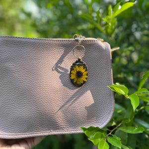 May include: A light mauve leather clutch with a sunflower keychain. The keychain features a yellow sunflower encased in a clear resin oval with a bronze-coloured frame. The clutch has a textured surface.