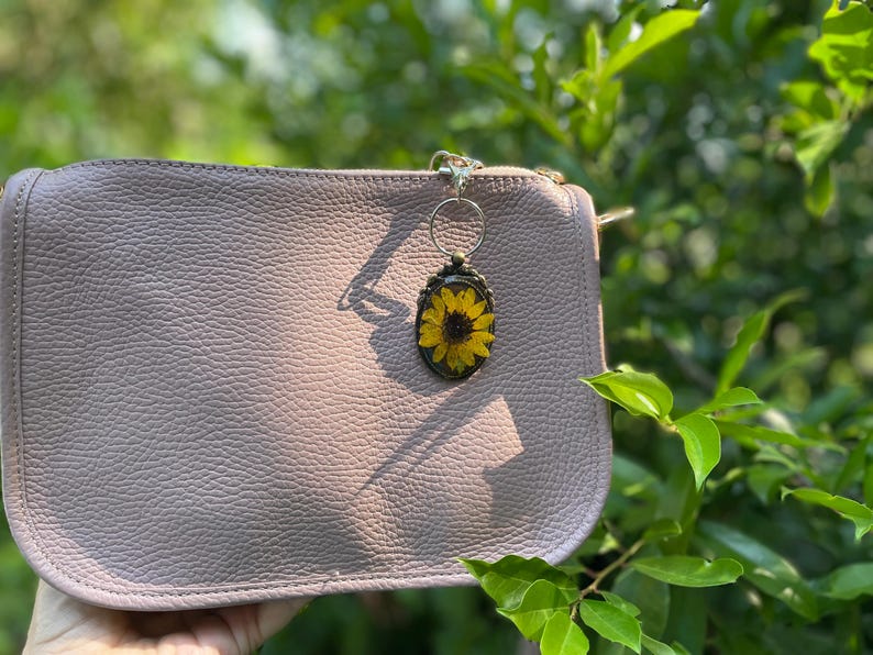 May include: A light mauve leather clutch with a sunflower keychain. The keychain features a yellow sunflower encased in a clear resin oval with a bronze-coloured frame. The clutch has a textured surface.