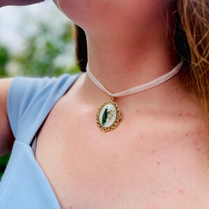 Pode incluir: Um colar de fita branca com um pingente oval dourado com uma flor de l&iacute;rio do vale prensada.