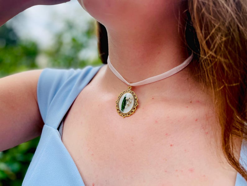 Pode incluir: Um colar de fita branca com um pingente oval dourado com uma flor de l&iacute;rio do vale prensada.