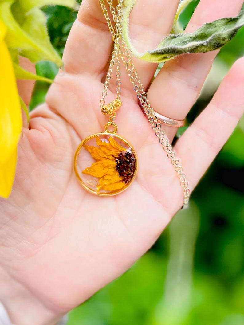 May include: A gold necklace with a circular pendant containing a preserved sunflower. The pendant showcases a yellow and brown sunflower, set in clear resin. The fine gold chain has a simple clasp, creating an elegant piece of jewellery.