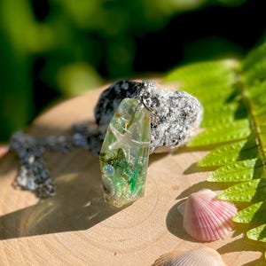 Op de afbeelding: Een heldere hars hanger met ingebedde schelpen, zeesterren en groen plantenleven, hangend aan een zilveren ketting. De hanger wordt gepresenteerd op een houten oppervlak met twee schelpen en een steen op de achtergrond. De hanger is ongeveer 5 cm lang.