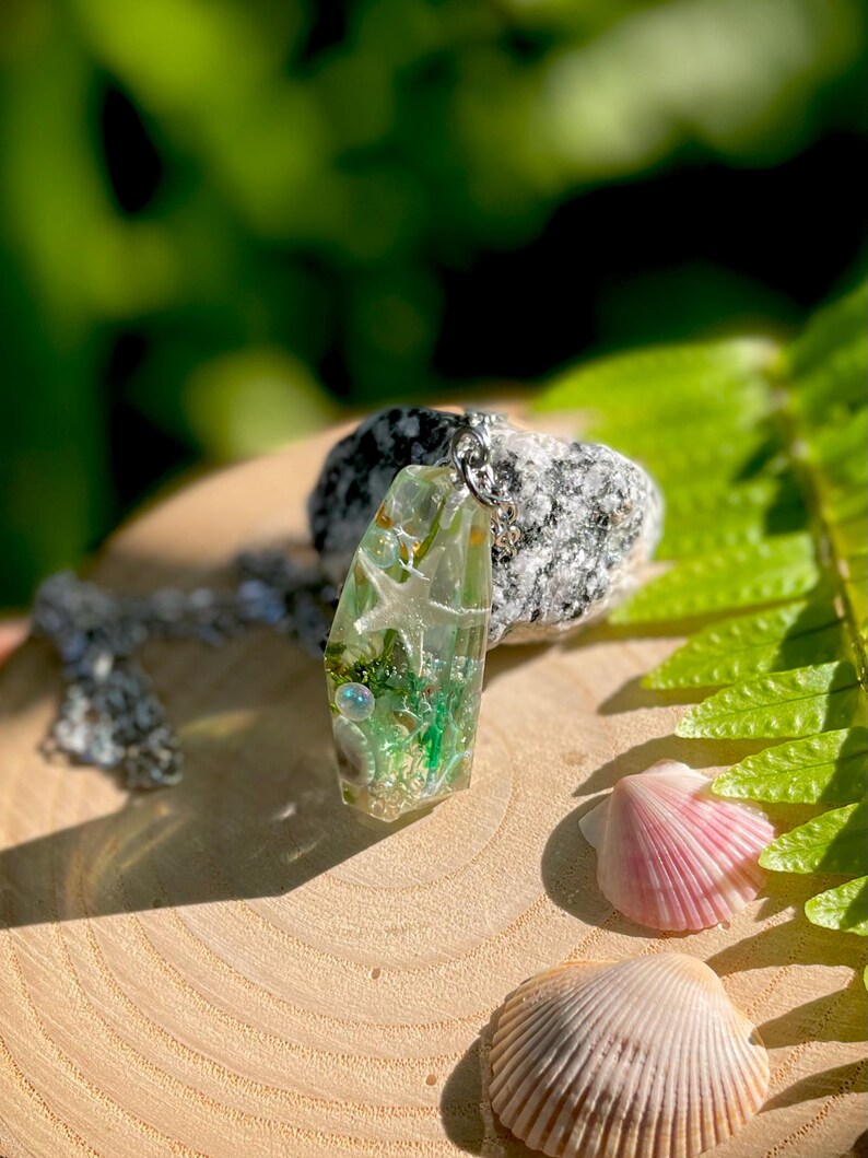 Op de afbeelding: Een heldere hars hanger met ingebedde schelpen, zeesterren en groen plantenleven, hangend aan een zilveren ketting. De hanger wordt gepresenteerd op een houten oppervlak met twee schelpen en een steen op de achtergrond. De hanger is ongeveer 5 cm lang.