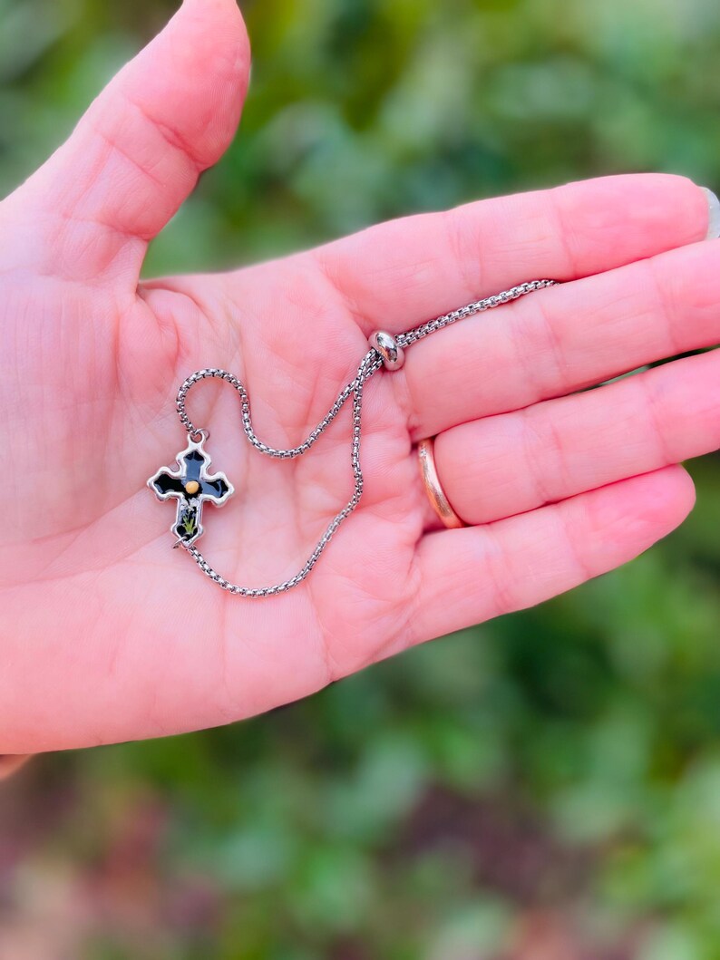 May include: A silver chain bracelet with a black cross charm featuring a gold center. The cross has a detailed design and is accented with small green stones.