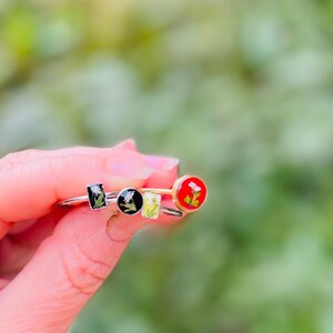 May include: Three silver rings with different colored resin insets. The insets feature pressed flowers. One ring has a red inset, one has a black inset, and one has a green inset.