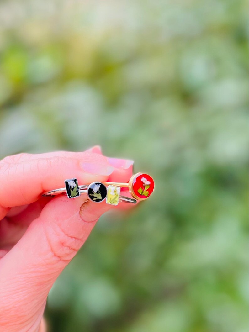 May include: Three silver rings with different colored resin insets. The insets feature pressed flowers. One ring has a red inset, one has a black inset, and one has a green inset.