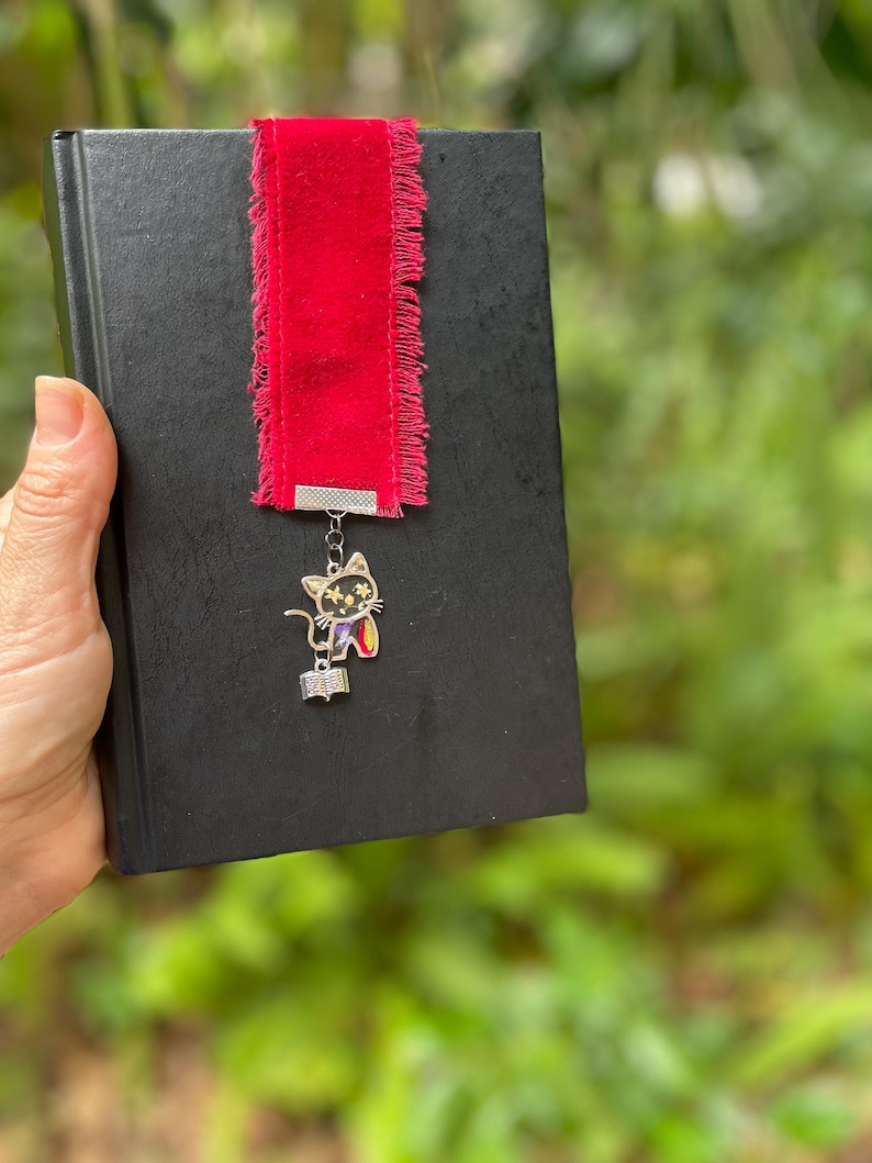 May include: A red velvet bookmark with frayed edges and a silver cat charm holding a book. The bookmark is attached to a black book.