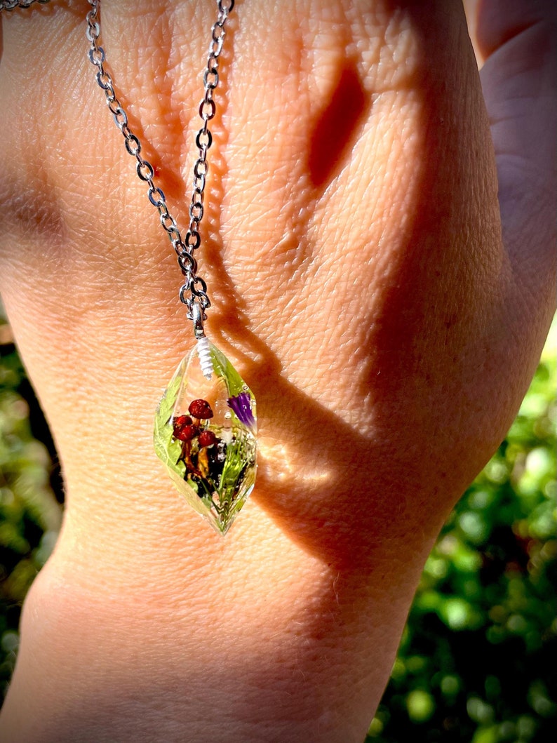 May include: A silver chain necklace with a clear teardrop-shaped pendant containing pressed red and purple flowers and green leaves.