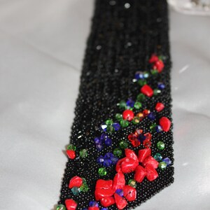 May include: Black beaded accessory with red, green, and blue accents. The accessory features a floral design with red stones and colorful beads. The background is a white, silky fabric.