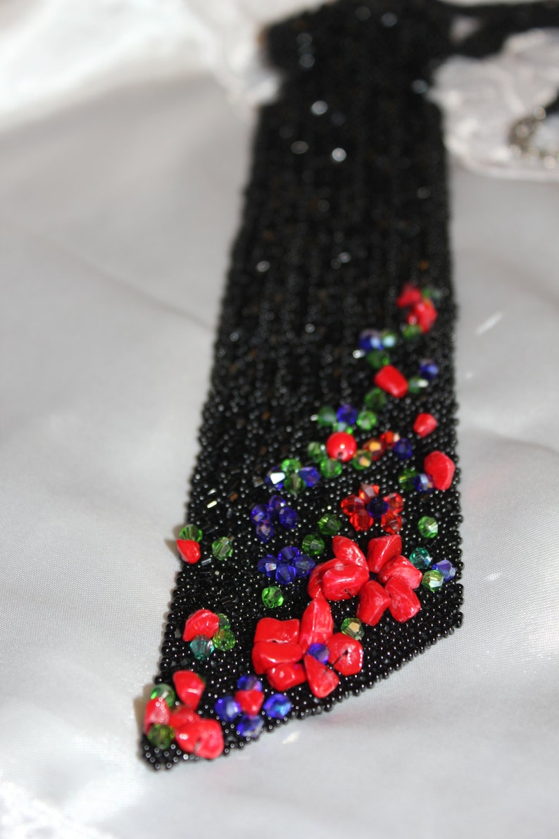 May include: Black beaded accessory with red, green, and blue accents. The accessory features a floral design with red stones and colorful beads. The background is a white, silky fabric.