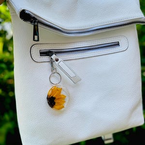 May include: White leather backpack with multiple zipped compartments. A clear resin keyring with a yellow sunflower is attached to the front zip. The backpack has a top flap and a handle.